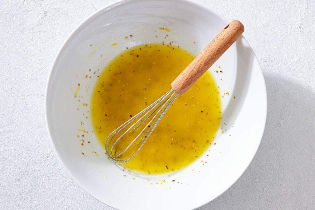 A metal whisk rests in a white bowl containing a yellow liquid vinaigrette with visible herbs.