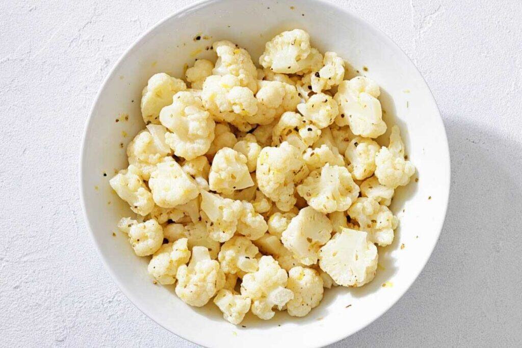 A white bowl filled with seasoned cauliflower florets on a light-colored surface.