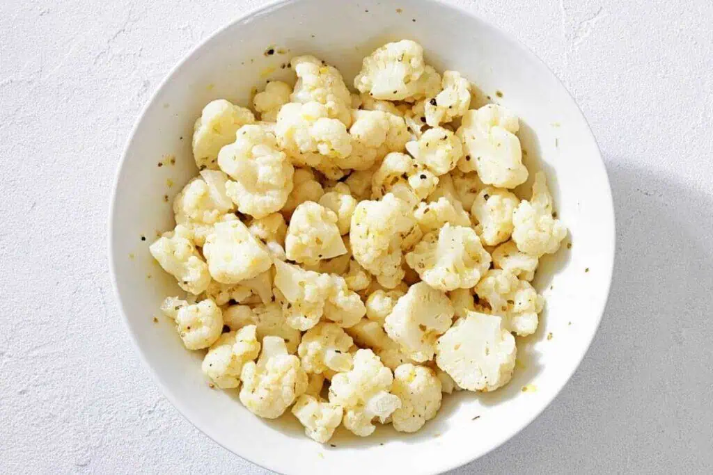 A white bowl filled with seasoned cauliflower florets on a light-colored surface.