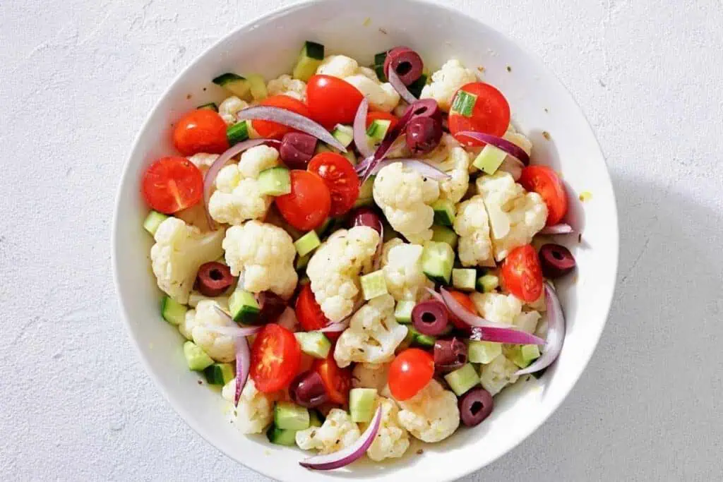 A bowl of cauliflower salad with cherry tomatoes, cucumber, black olives, red onion, and a light dressing on a white background.