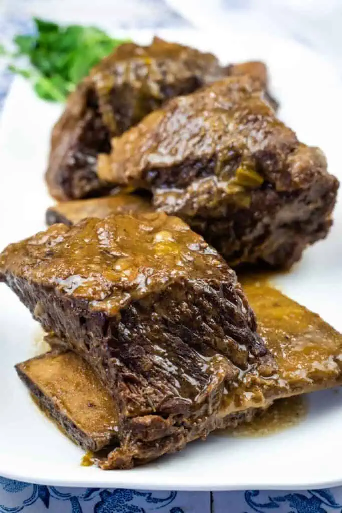 Three pieces of braised beef short ribs with sauce are served on a white plate, with some greenery in the background.