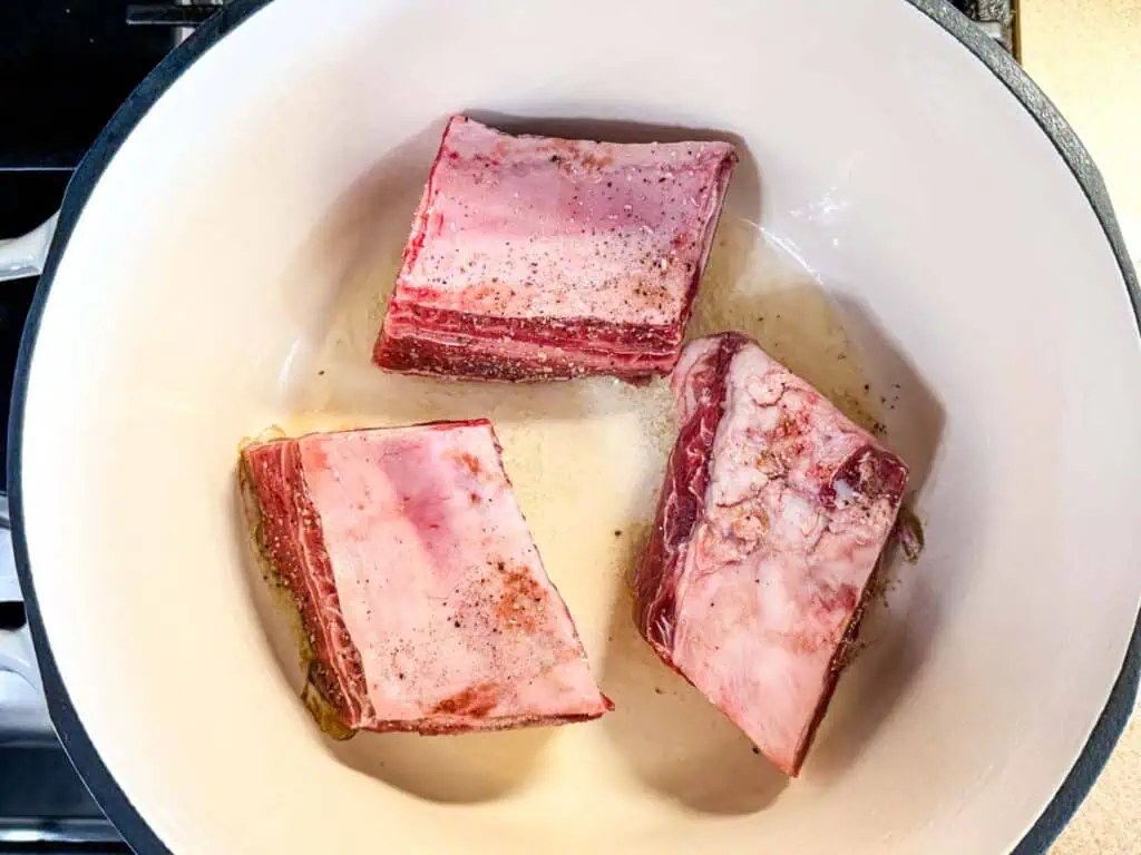 Three raw, seasoned beef short ribs are browning in a white Dutch oven on a stovetop.