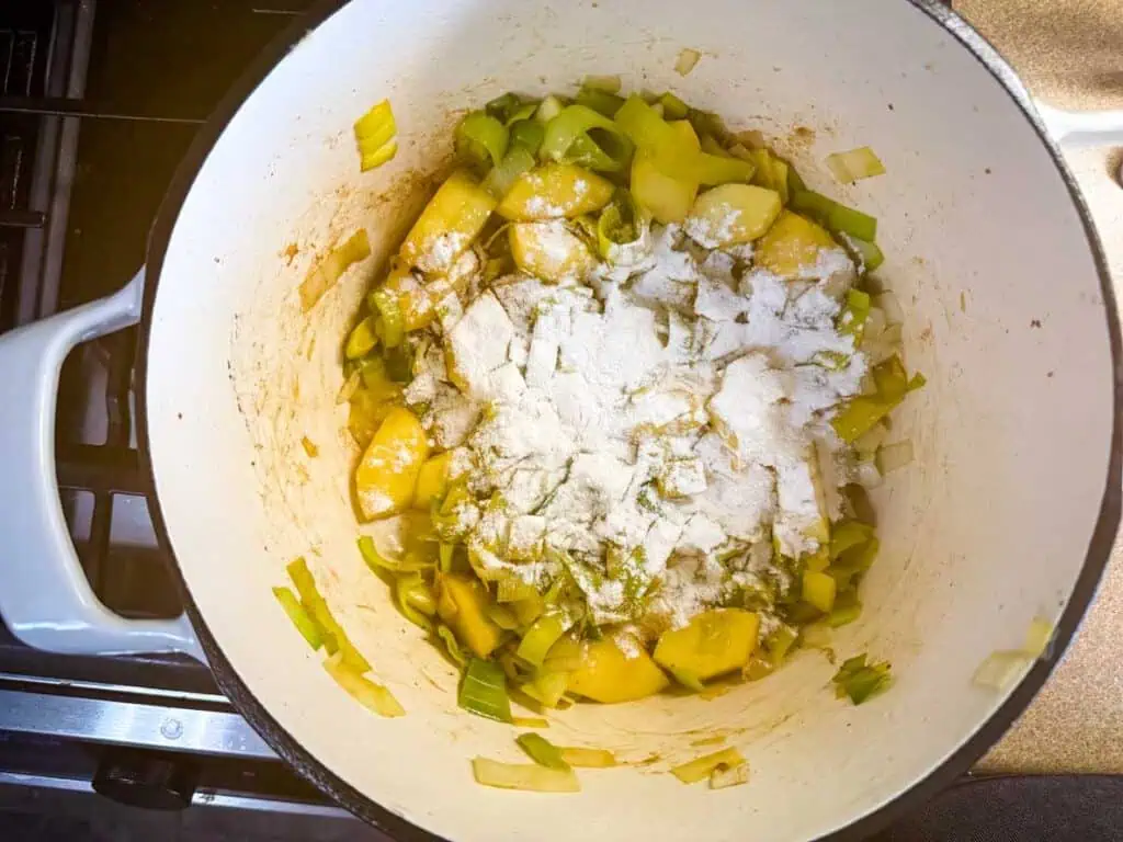 Chopped leeks, potatoes, and flour are in a white pot on a stove, ready for cooking.