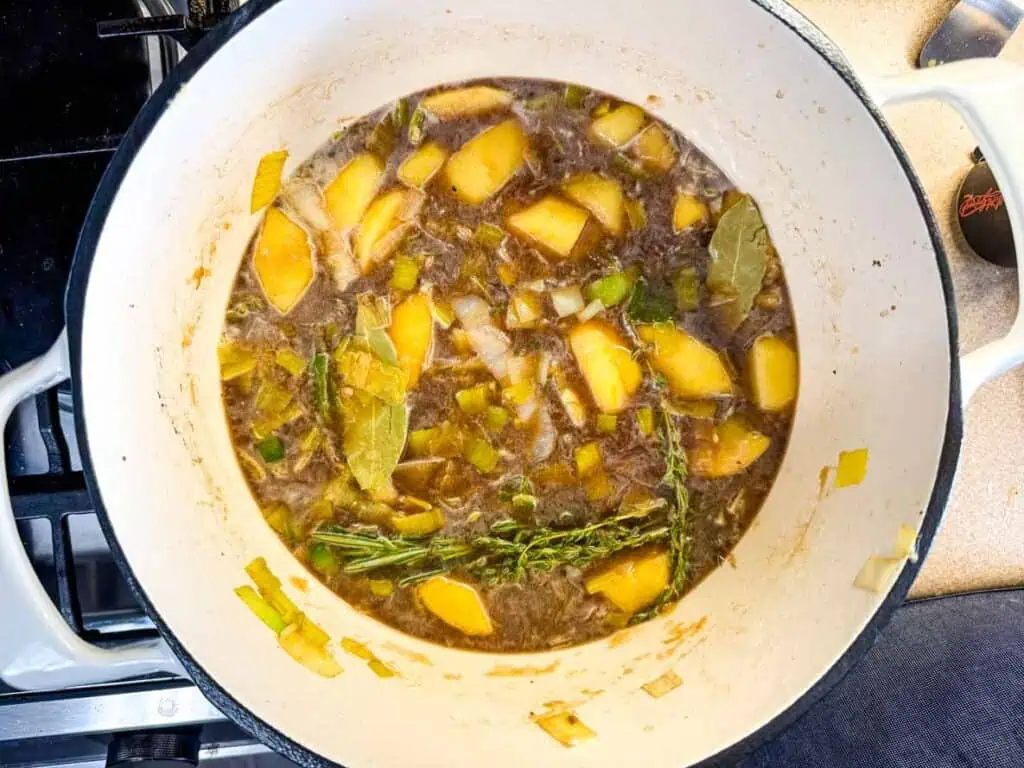 A white pot on a stove contains a simmering mixture of chopped potatoes, leeks, thyme, bay leaves, and broth.