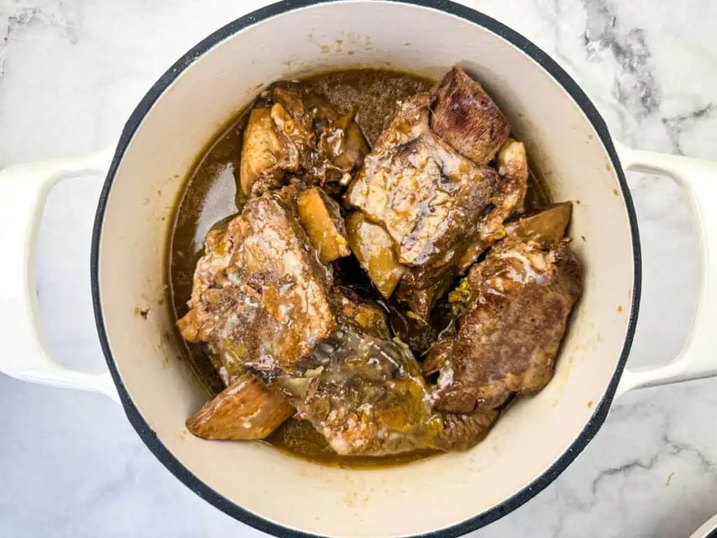 Cooked beef short ribs in sauce inside a white Dutch oven, set on a marble countertop.
