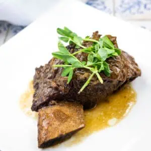 A cooked beef short rib with sauce, garnished with fresh greens, served on a white plate.