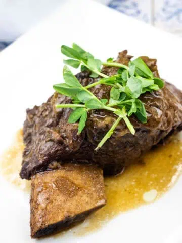 A cooked beef short rib with sauce, garnished with fresh greens, served on a white plate.
