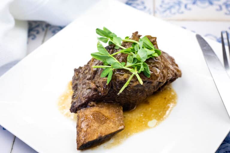 A cooked beef short rib with sauce, garnished with fresh greens, served on a white plate.