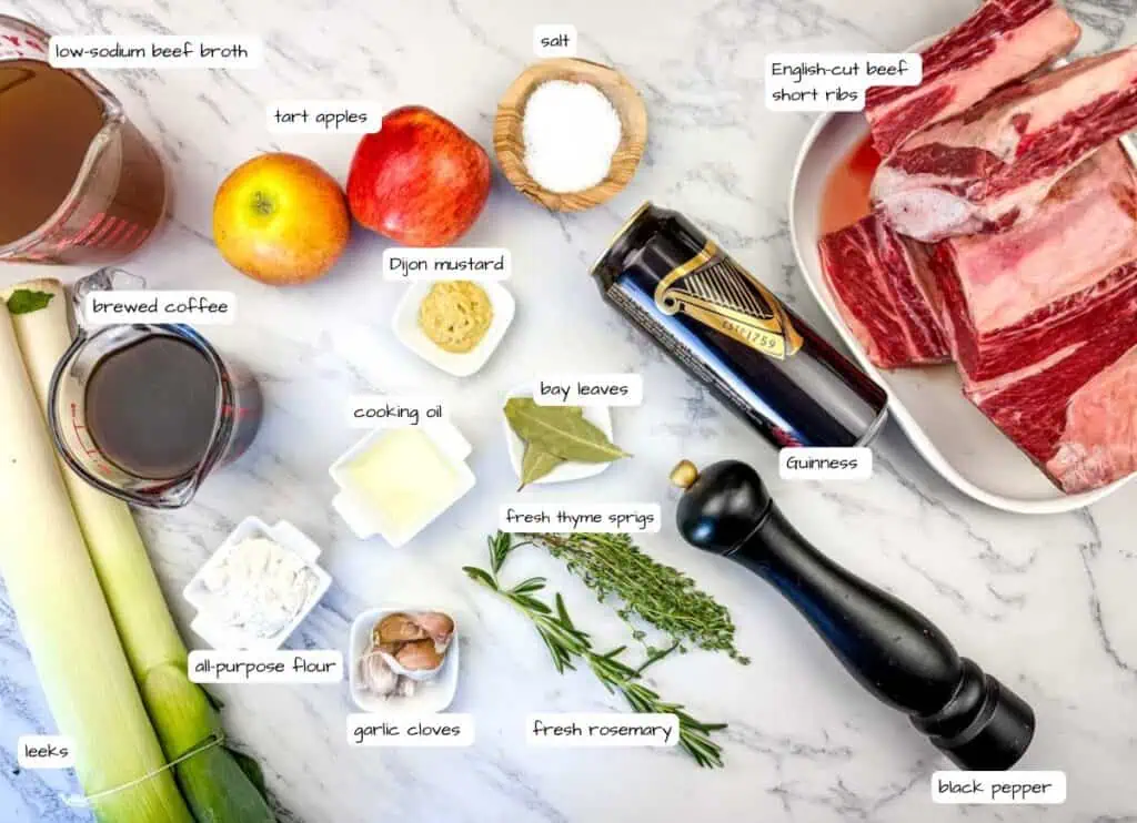 A variety of labeled ingredients for braised beef short ribs are arranged on a marble countertop, including apples, leeks, herbs, spices, and liquids.