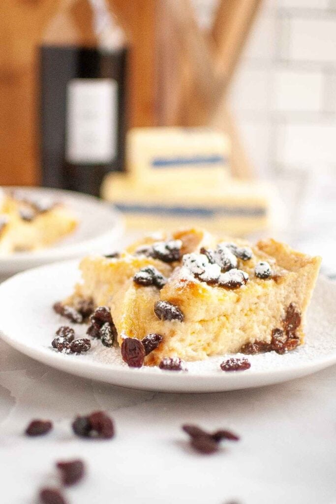 A slice of bread pudding topped with raisins and powdered sugar is served on a white plate, with more bread pudding and blurred ingredients in the background.