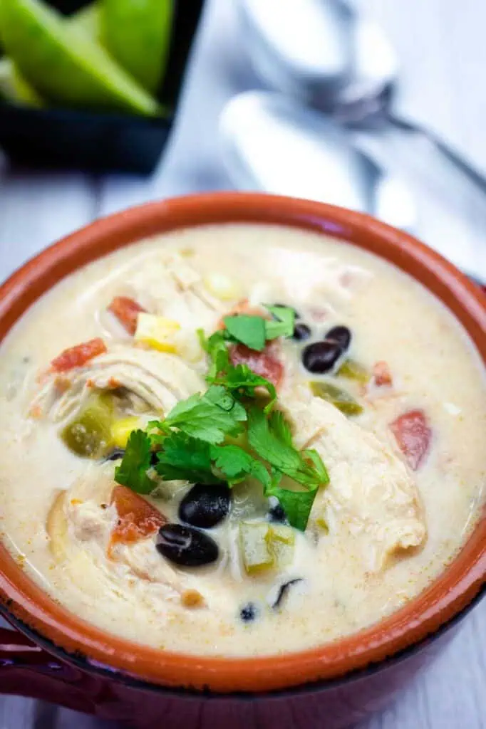 A bowl of creamy chicken soup with black beans, tomatoes, green peppers, and fresh cilantro garnish, with spoons and a small dish of lime wedges in the background.