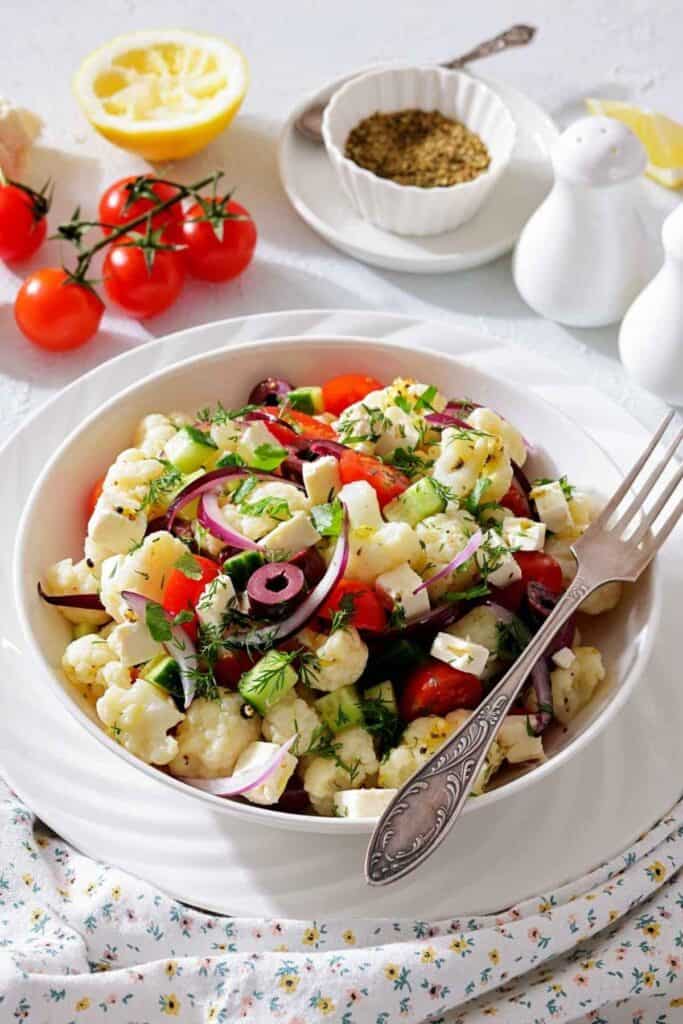 A bowl of cauliflower salad with tomatoes, olives, red onions, feta cheese, and fresh herbs, set on a white plate with a fork; salt, pepper, and seasoning in the background.