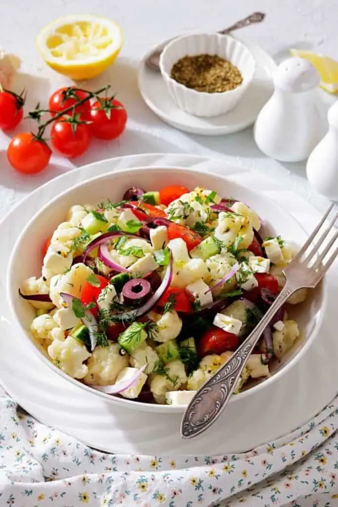 A bowl of cauliflower salad with tomatoes, olives, red onions, feta cheese, and fresh herbs, set on a white plate with a fork; salt, pepper, and seasoning in the background.