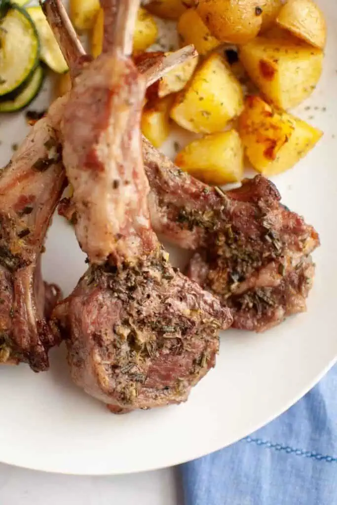 Three cooked lamb chops garnished with herbs on a white plate, served with roasted potato cubes and sliced zucchini.