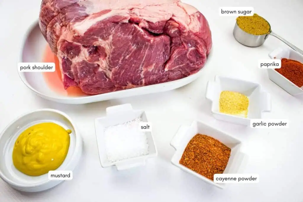 A raw pork shoulder on a white tray is surrounded by small dishes of mustard, salt, brown sugar, paprika, garlic powder, and cayenne powder, each labeled.