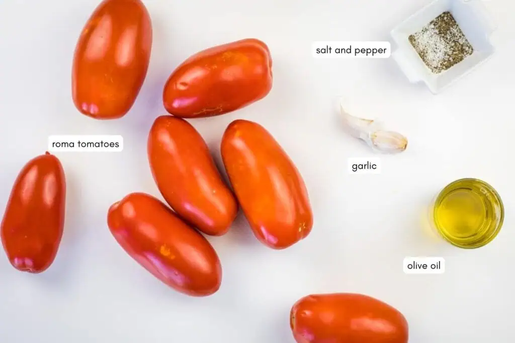 Six Roma tomatoes, two garlic cloves, a dish of salt and pepper, and a small container of olive oil arranged on a white surface.