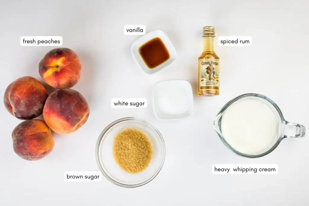A flat lay of fresh peaches, brown sugar, white sugar, vanilla, spiced rum, and heavy whipping cream on a white surface, each ingredient labeled.
