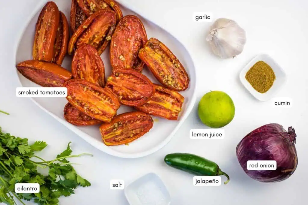A tray of smoked tomato halves is surrounded by cilantro, salt, a jalapeño, a lime, a red onion, garlic, and a small bowl of cumin. Each item is labeled.