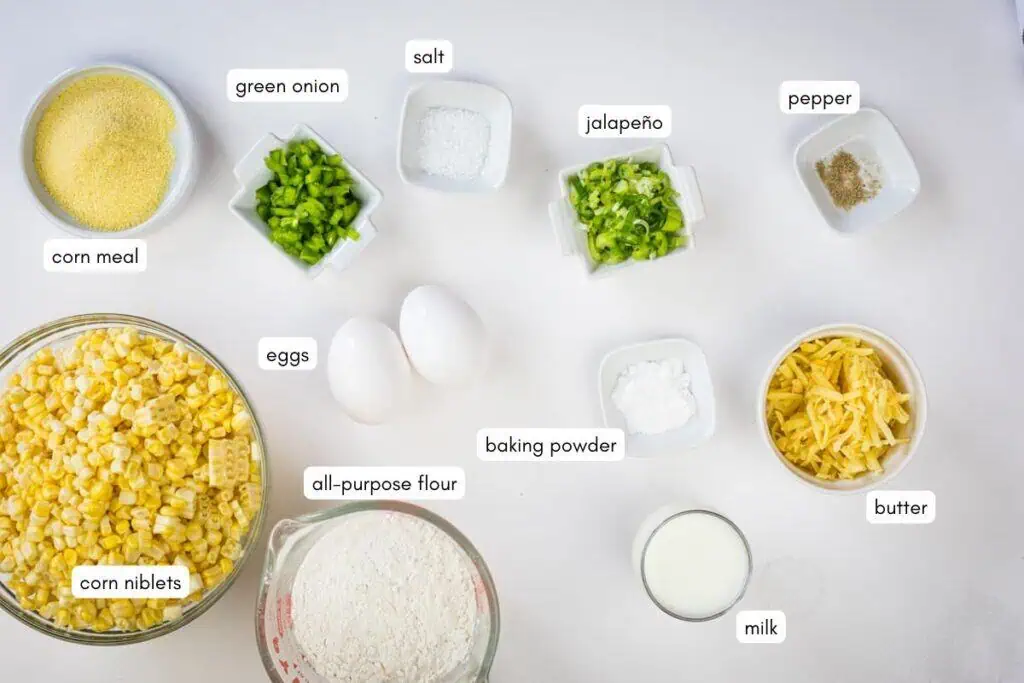 Ingredients for cornbread arranged on a white surface, including corn meal, corn niblets, green onion, eggs, flour, salt, jalapeño, baking powder, cheese, butter, pepper, and milk.
