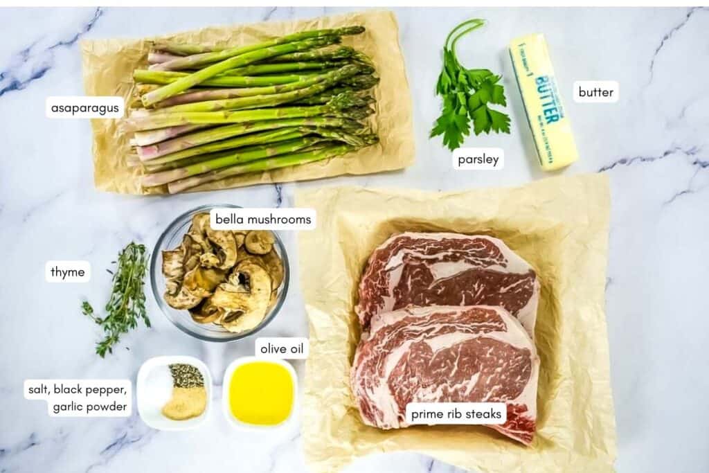Ingredients on a marble surface: asparagus, parsley, butter, prime rib steaks, olive oil, bella mushrooms, thyme, and small bowls of salt, black pepper, and garlic powder.