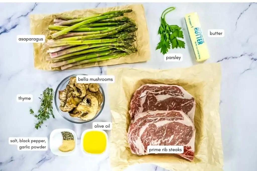 Ingredients on a marble surface: asparagus, parsley, butter, prime rib steaks, olive oil, bella mushrooms, thyme, and small bowls of salt, black pepper, and garlic powder.