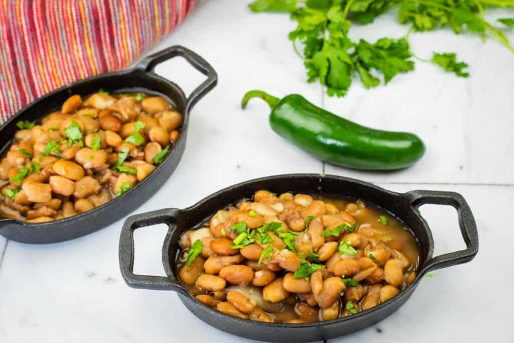 Two black oval dishes filled with Charro Beans, garnished with cilantro. A whole jalape&ntilde;o and cilantro sprigs are on a marble surface nearby.