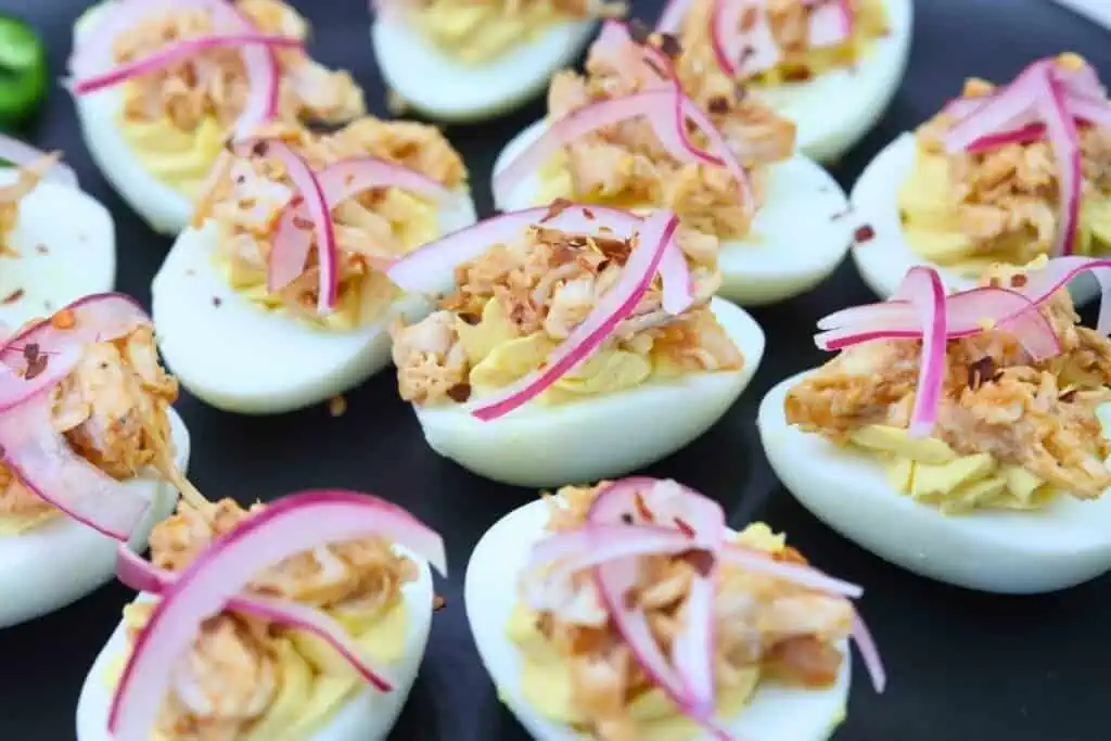Deviled eggs topped with shredded meat and thinly sliced red onions are arranged on a black plate.