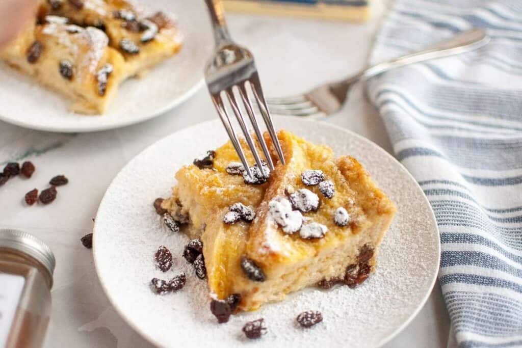 A plate with two slices of bread pudding topped with powdered sugar and raisins.