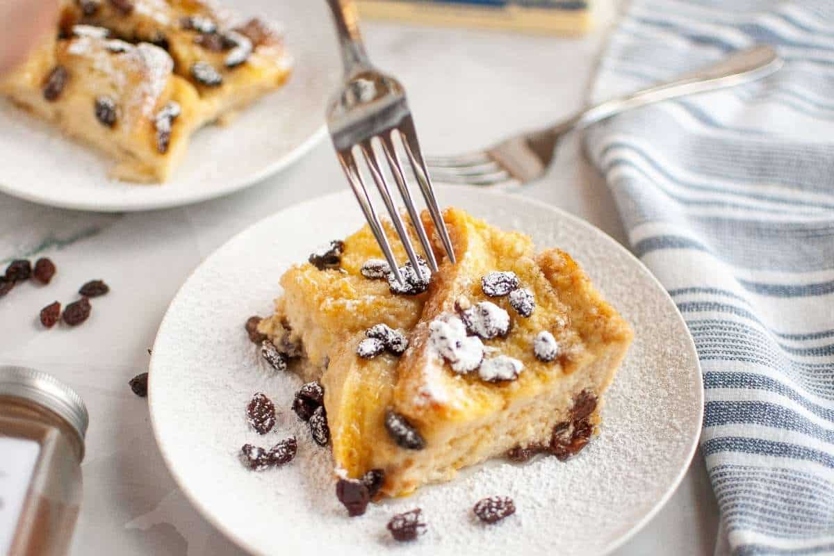 A fork cutting into a slice of bread pudding with raisins, dusted with powdered sugar, served on a white plate.