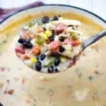 A ladle holds a serving of creamy soup with black beans, corn, tomatoes, green peppers, and shredded chicken over a pot of the same soup.