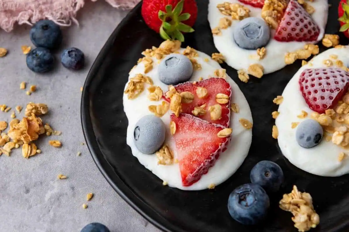 Three Frozen Vanilla Yogurt Bites topped with sliced strawberries, blueberries, and granola are arranged on a black plate, with extra berries and granola scattered around on the gray surface.