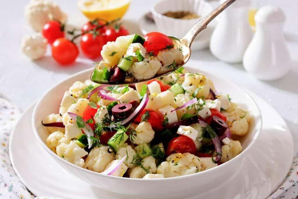 A bowl of cauliflower salad with cherry tomatoes, cucumbers, red onions, feta cheese, and herbs, with a spoon serving a portion.