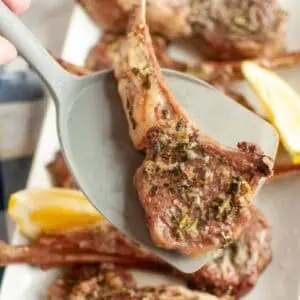 A hand holding a spatula lifts a seasoned, cooked lamb chop from a platter with more lamb chops and lemon wedges.