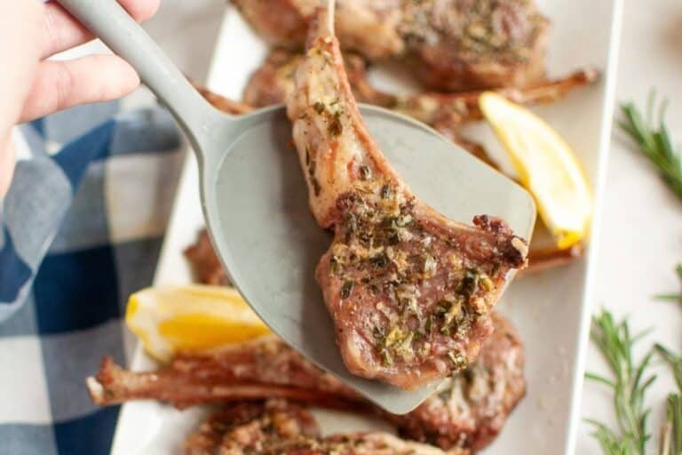 A hand holding a spatula lifts a seasoned, cooked lamb chop from a platter with more lamb chops and lemon wedges.