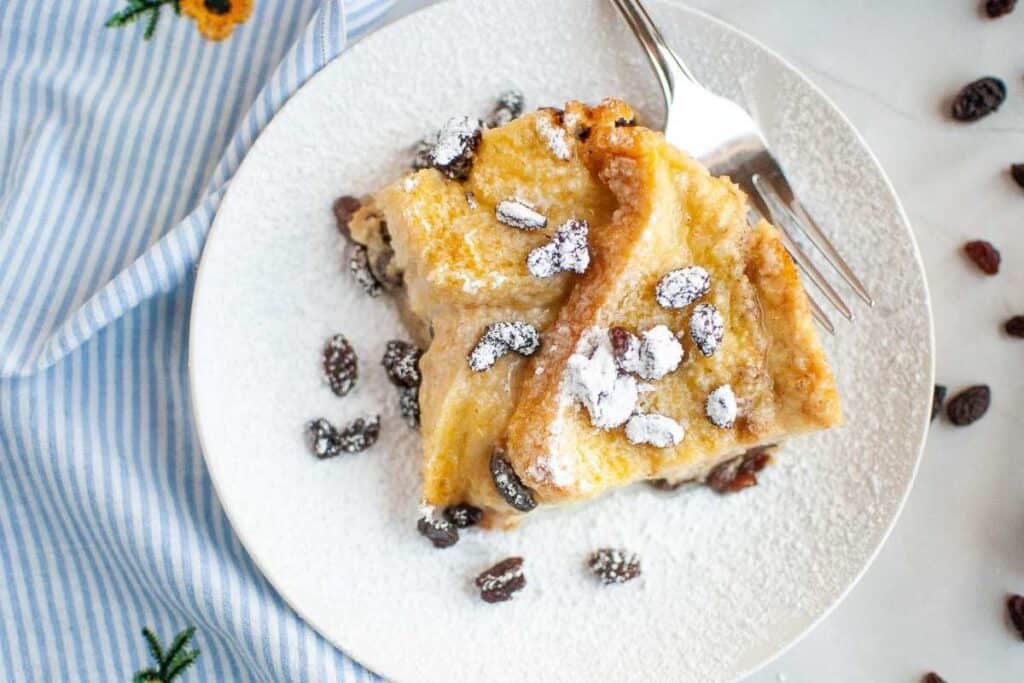 A plate with two slices of bread pudding topped with powdered sugar and raisins, next to a fork on a white surface.