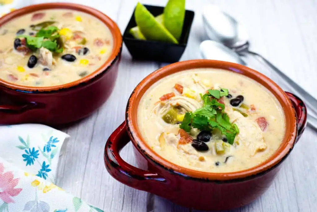 Two bowls of creamy soup with chicken, black beans, corn, and vegetables, garnished with cilantro. Lime wedges and spoons are in the background.