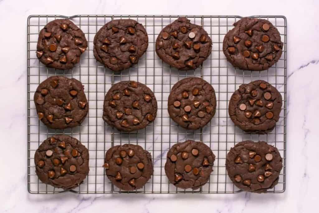 Twelve chocolate cookies with chocolate chips are cooling on a wire rack, placed on a marble surface.