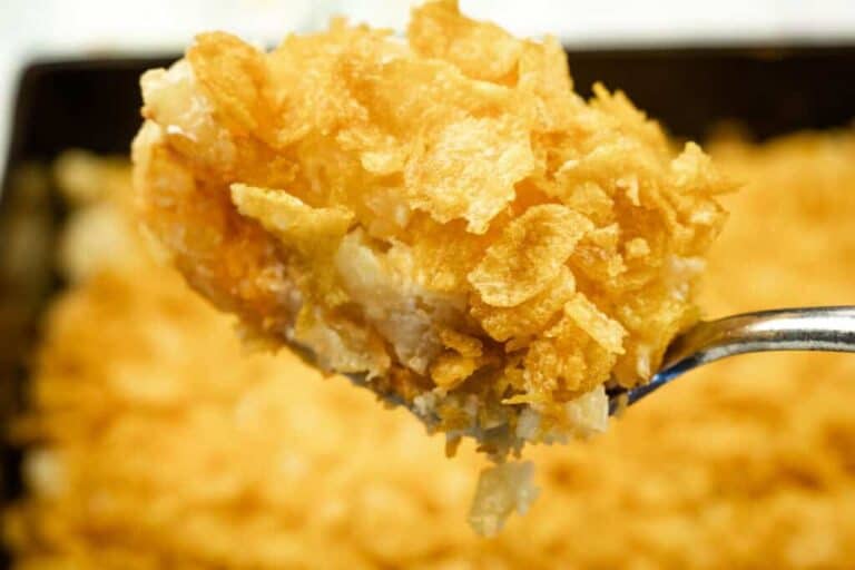 A close-up of a spoonful of casserole topped with a golden, crispy cornflake crust, with the rest of the casserole visible in the background.