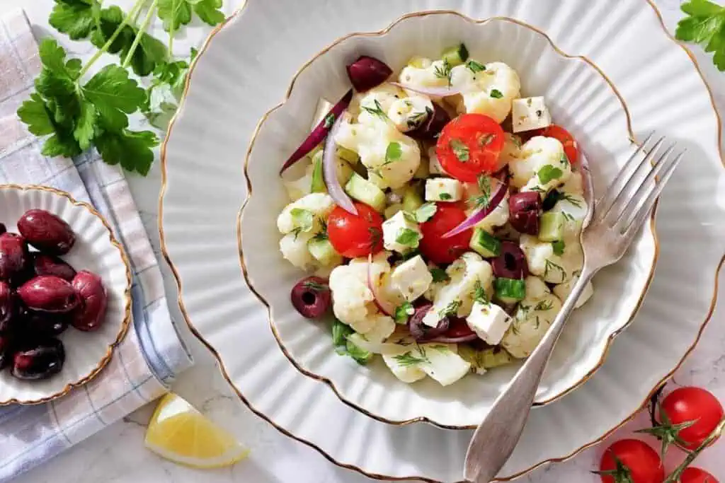 A plate of cauliflower salad with cherry tomatoes, olives, red onion, feta cheese, and herbs next to a fork; more olives and garnishes are on the side.