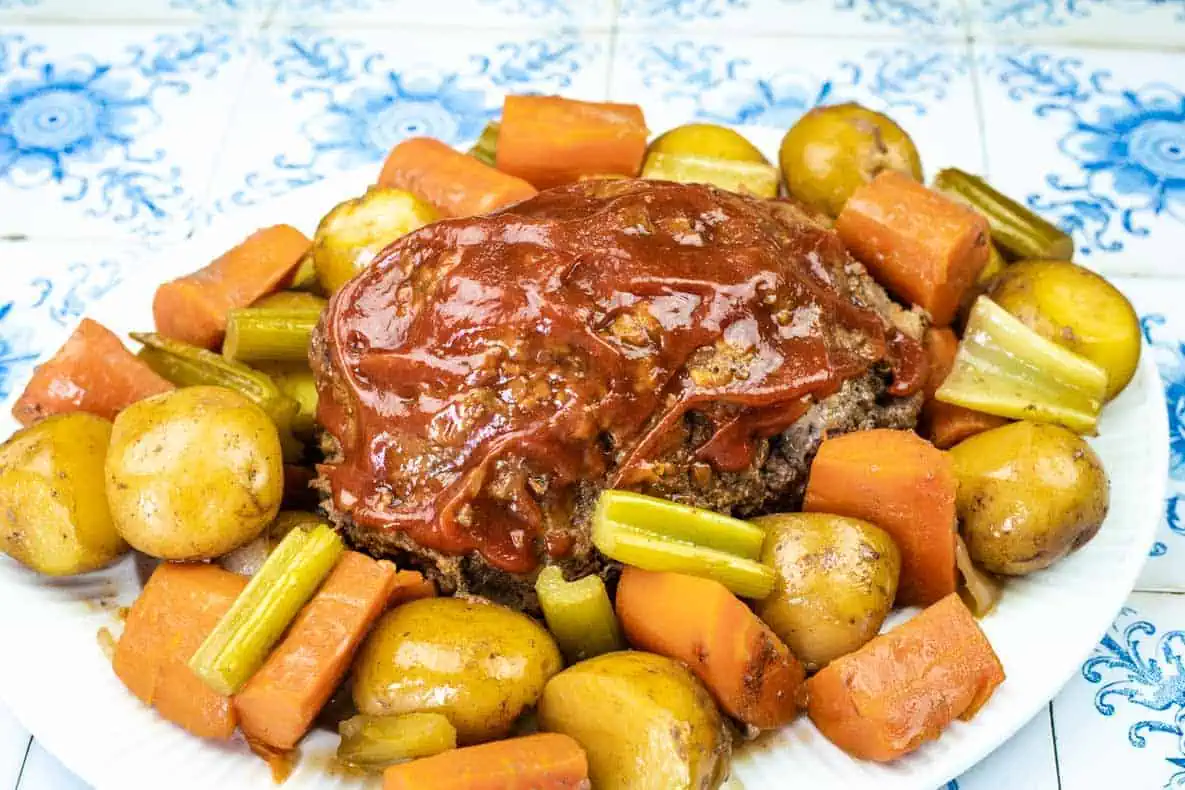 A plate of meatloaf topped with tomato sauce, surrounded by cooked baby potatoes, carrots, and celery on a patterned tile background.