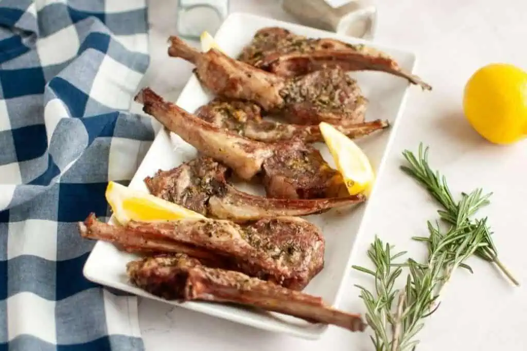 A rectangular plate with grilled lamb chops garnished with lemon wedges and herbs, alongside fresh rosemary and a blue checkered cloth.