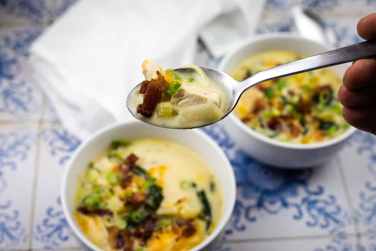 A hand holds a spoonful of Million dollar soup with chicken, bacon, and green onions above a bowl of soup on a blue and white tiled surface.