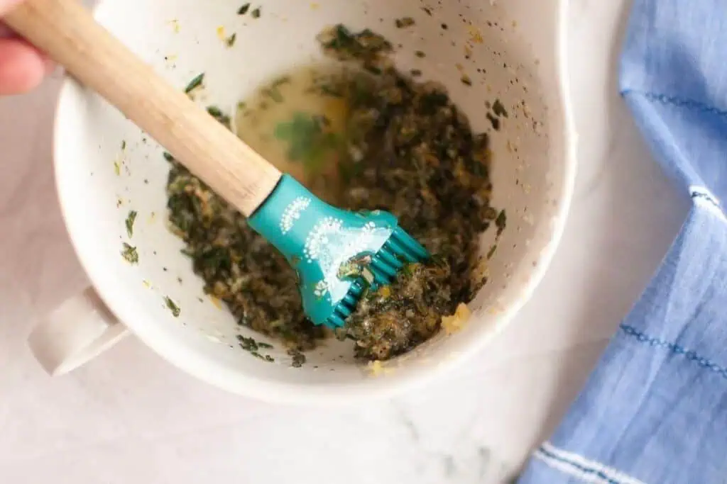A silicone basting brush rests in a white bowl with a mixture of chopped herbs and liquid, next to a blue cloth on a white surface.