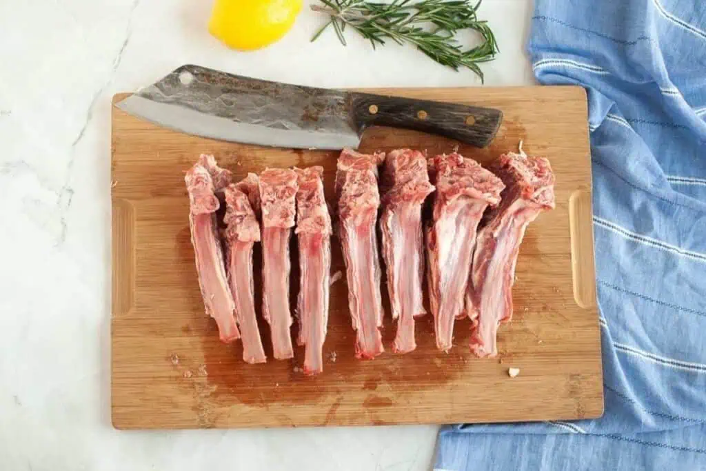 Raw lamb chops arranged in a row on a wooden cutting board with a large cleaver, rosemary, a lemon, and a blue cloth nearby.