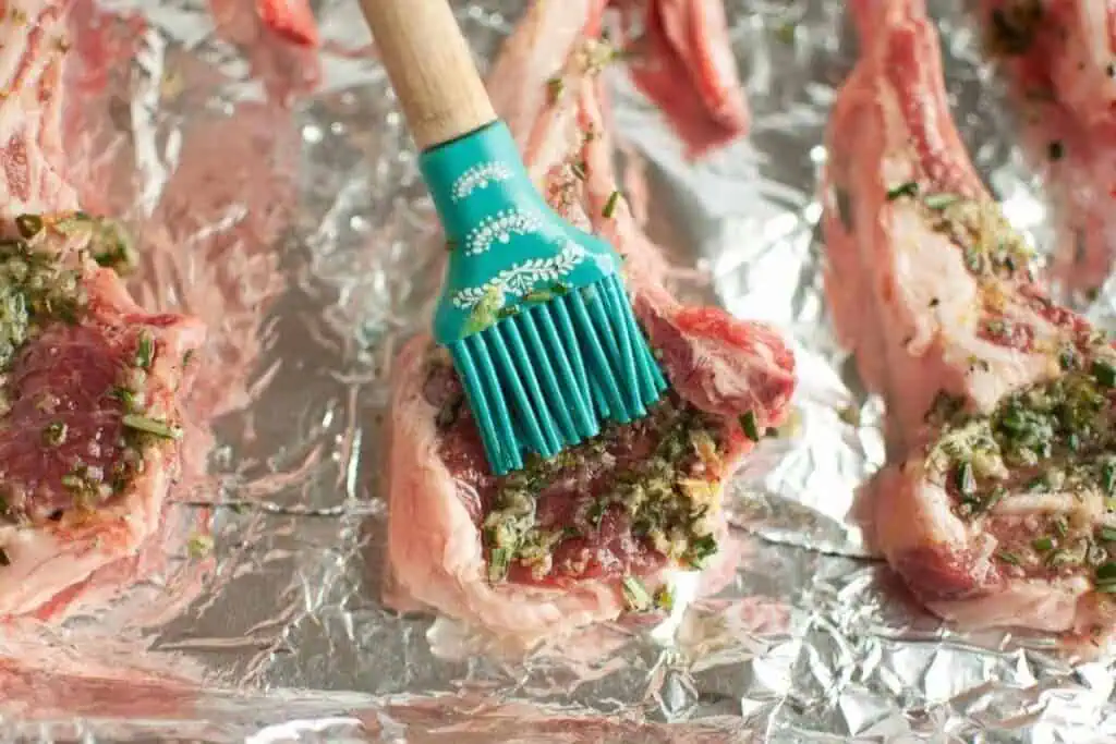 A silicone brush applies an herb marinade to raw lamb chops arranged on aluminum foil.