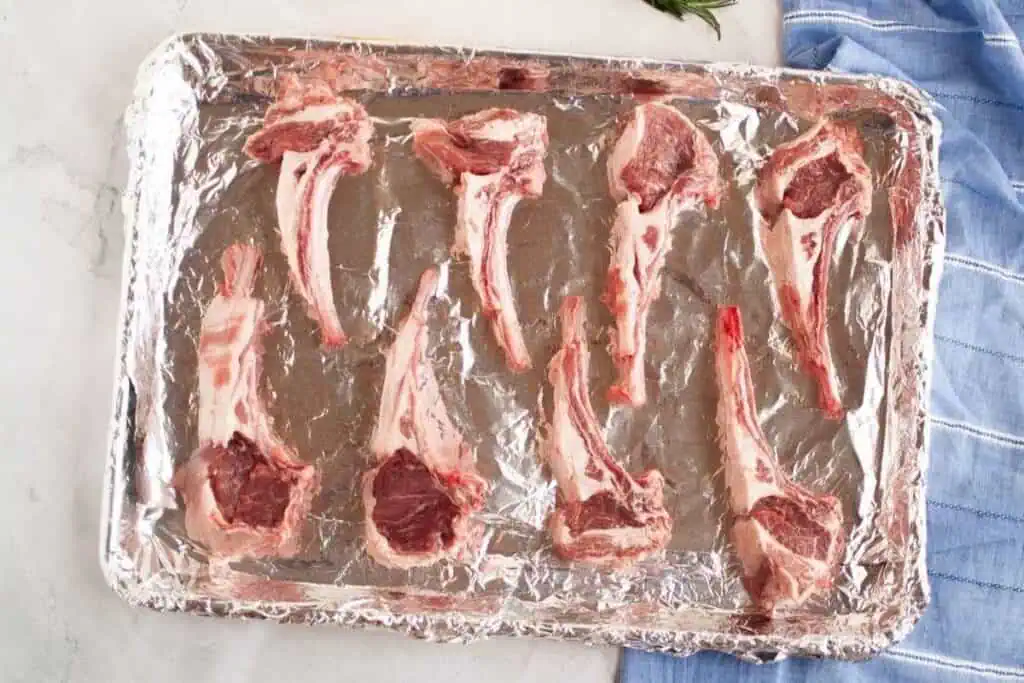 Eight raw lamb chops arranged on a foil-lined baking sheet, placed on a light-colored surface next to a blue striped cloth.