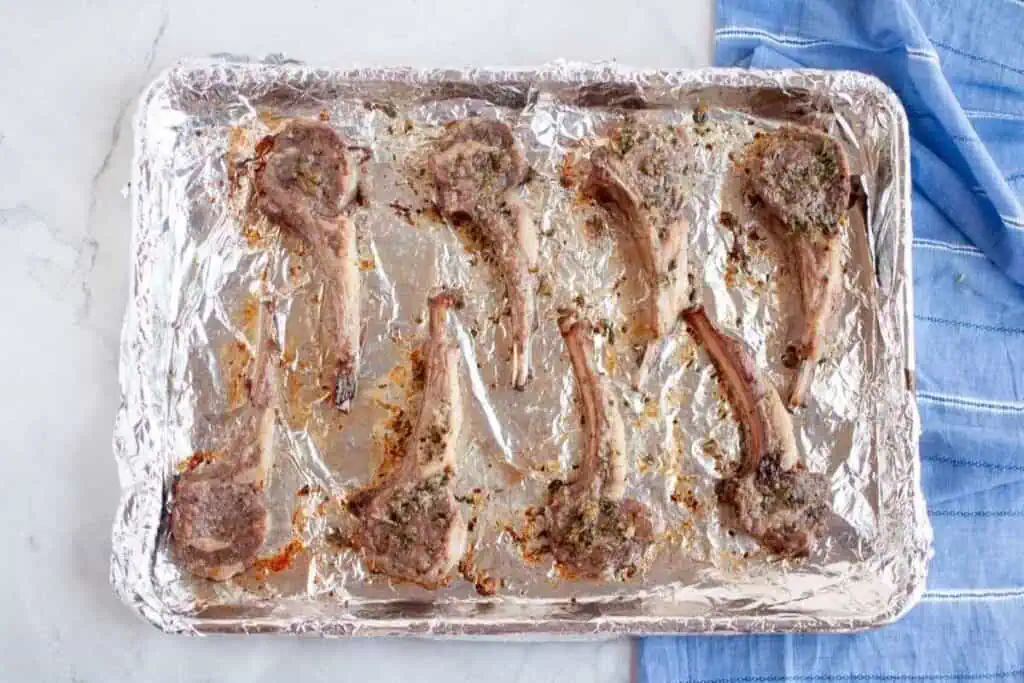 Eight cooked lamb chops arranged on a foil-lined baking sheet, resting on a light countertop next to a blue striped towel.