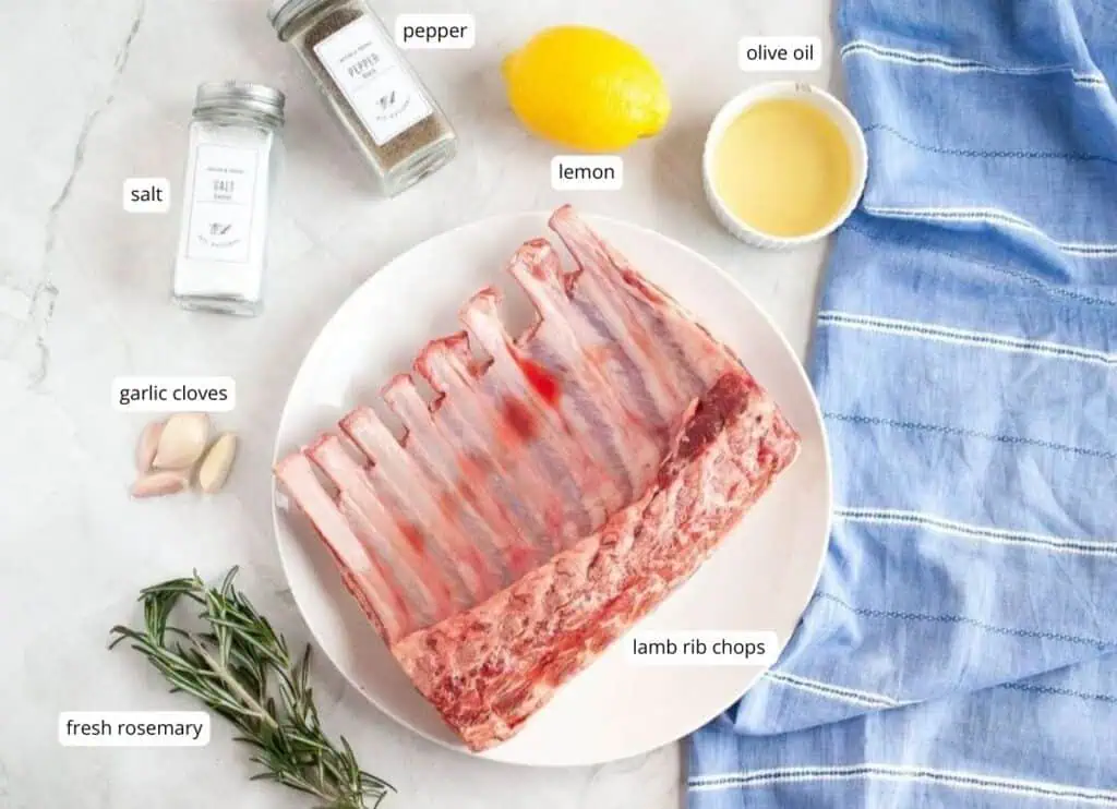 A plate of raw lamb rib chops is surrounded by fresh rosemary, garlic cloves, salt, pepper, a lemon, and a bowl of olive oil on a blue cloth. Each item is labeled.