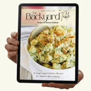Hands holding a tablet displaying the cover of "The Backyard Table" cookbook, featuring a bowl of potato salad and text about recipes for summer cookouts.