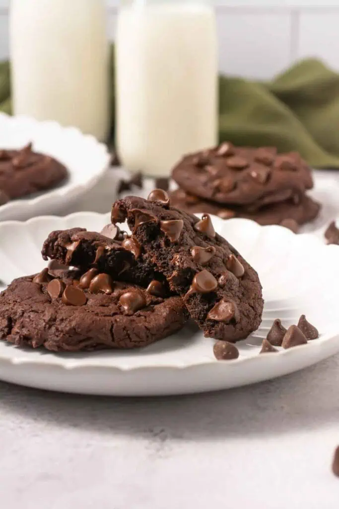 A plate of chocolate cookies with chocolate chips, one broken in half, sits in front of two glasses of milk.